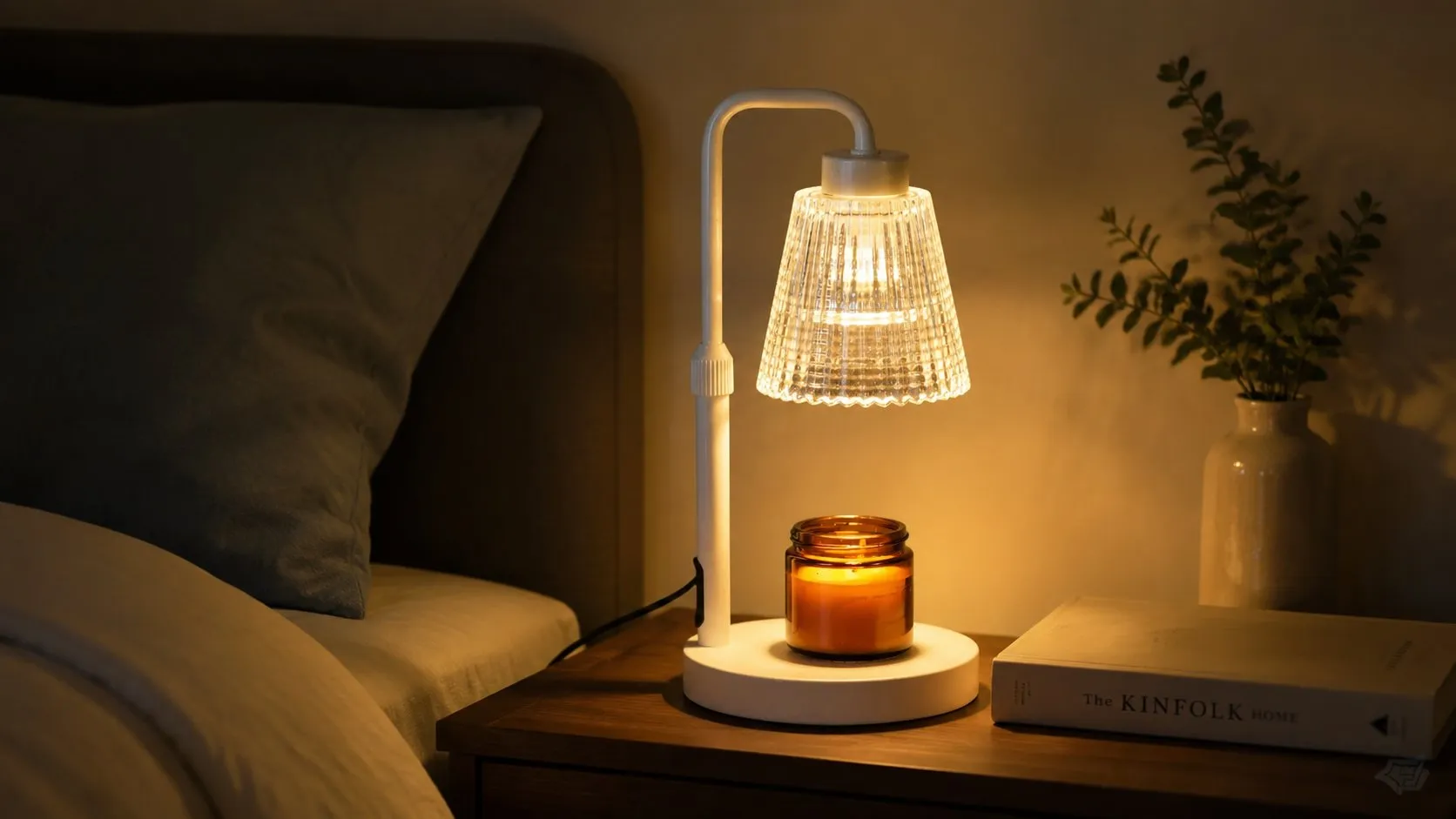 Ivory-white candle warmer lamp glowing on a bedside table at night beside a jar candle and a book, showing the flameless ambient-light use case shoppers buy for