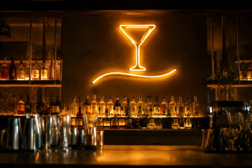 Warm amber neon cocktail glass sign glowing on a dark back bar wall above lit bottle shelving in a dim bar