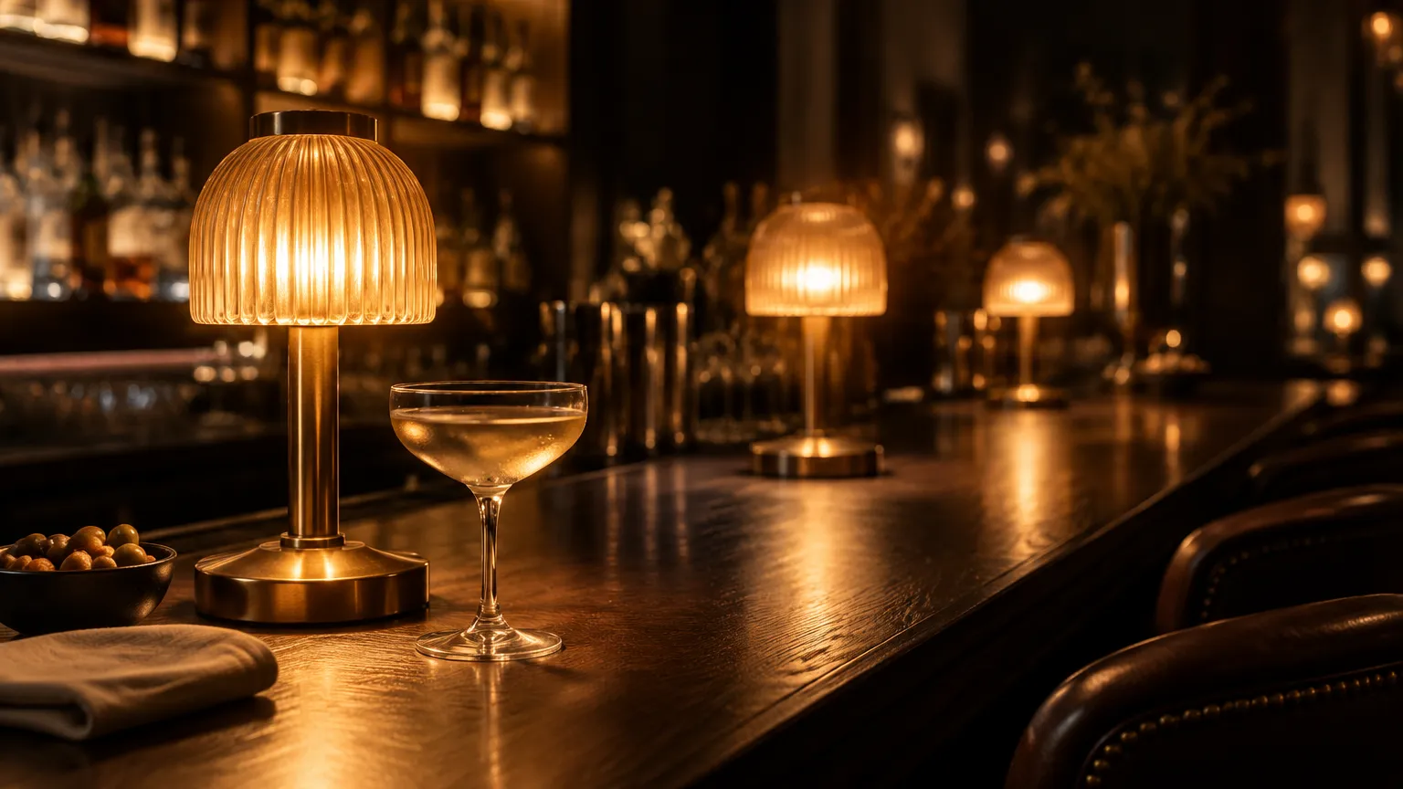 Row of small warm table lamps with fluted glass shades and aged brass bases along a dark timber bar top at night, glassware and stools in soft focus behind
