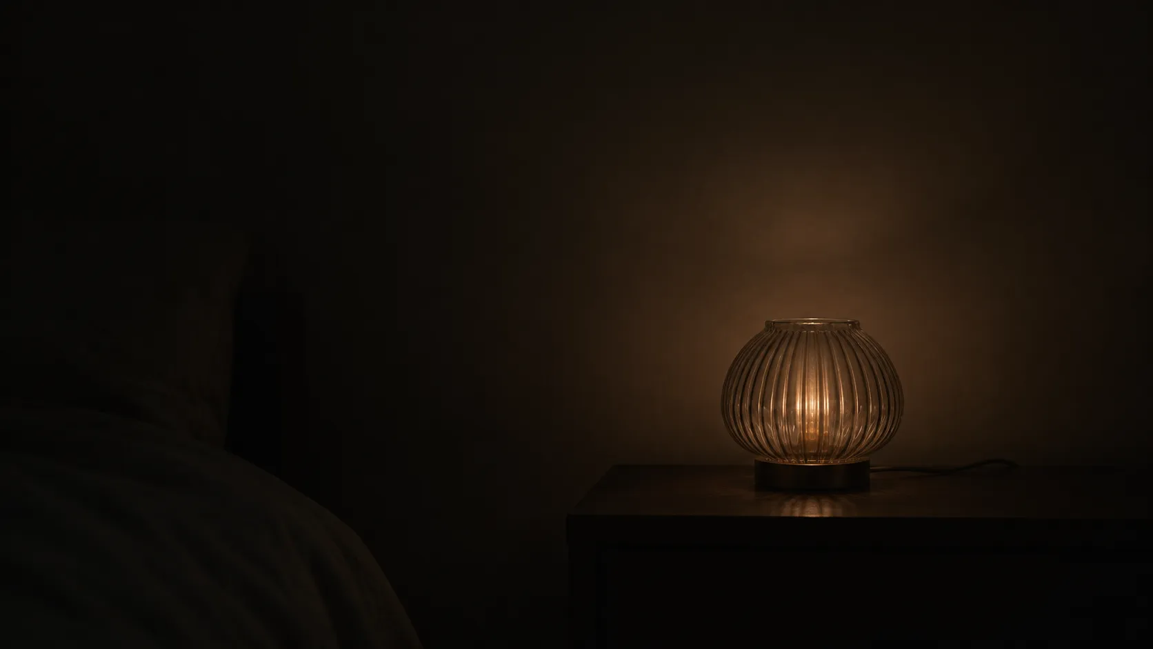 Decorative glass lamp at its lowest setting in a near-dark bedroom, a faint glow on the wall behind it