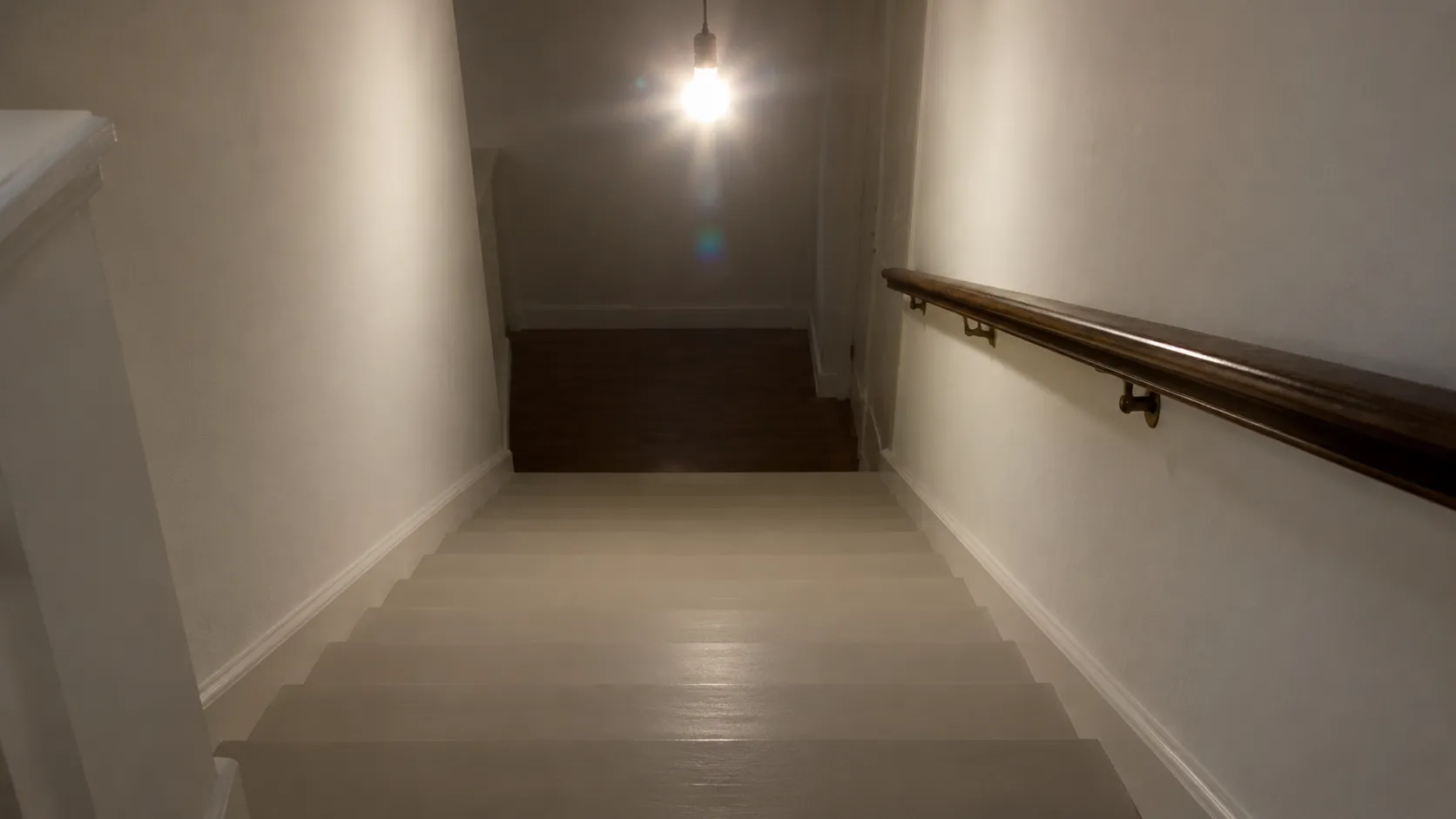 View down a wooden staircase from the top landing under one bright bare bulb, the step edges blending together